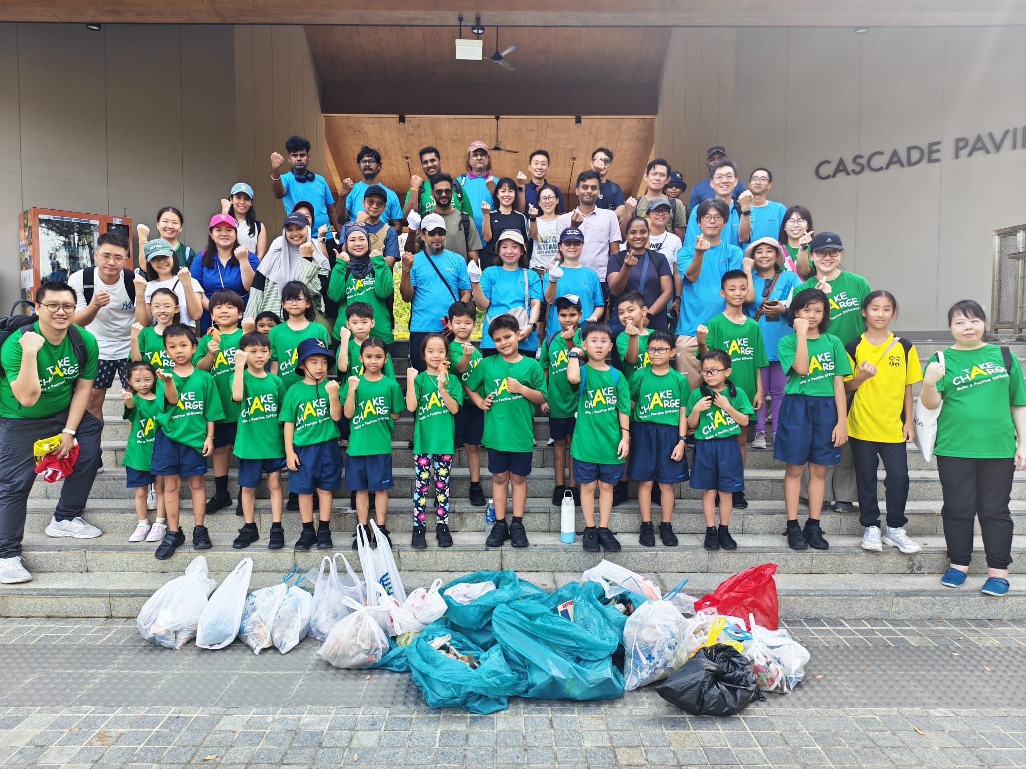 Our school community in green "Take Charge" shirts and PacificLight representatives pose on steps with collected trash bags in front